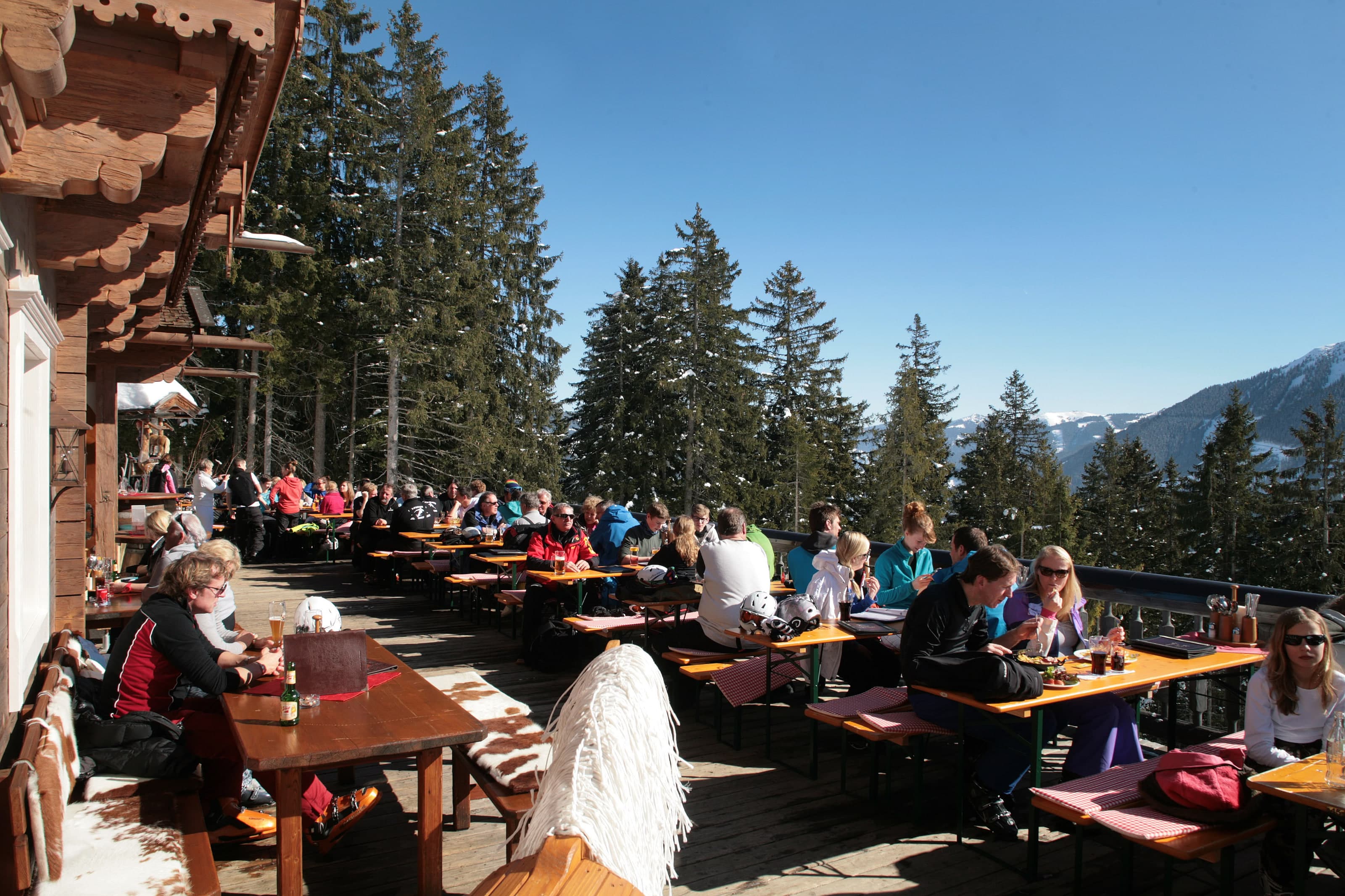 Gut besuchte Sonnenterrasse des Westernstadl, Holzbänke in der Sonne, dahinter Wald und verschneite Berge